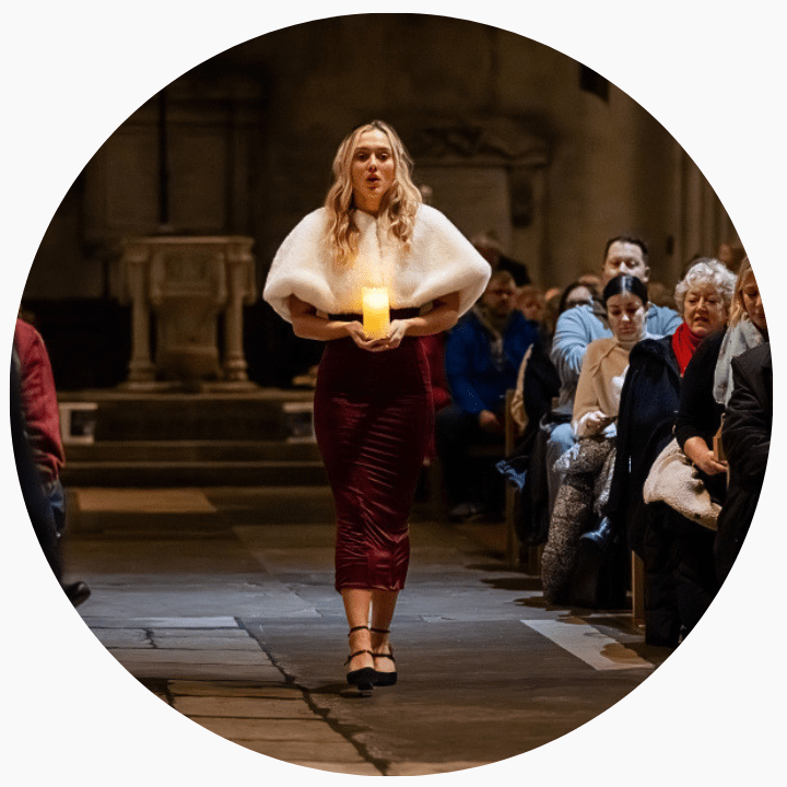 A woman in a red dress, with a white coat, walking down an aisle with a candle in her hand.