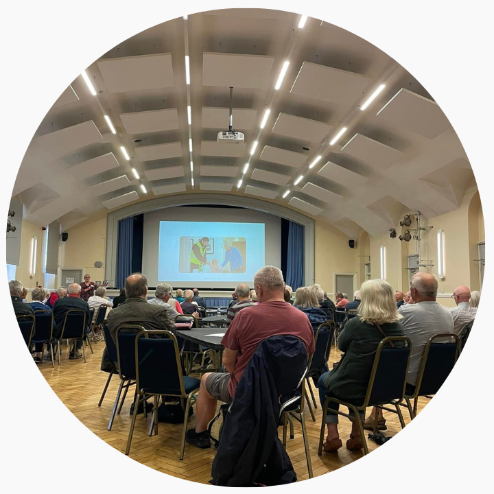 A crowd full of people sat in a room with tables and chairs.
